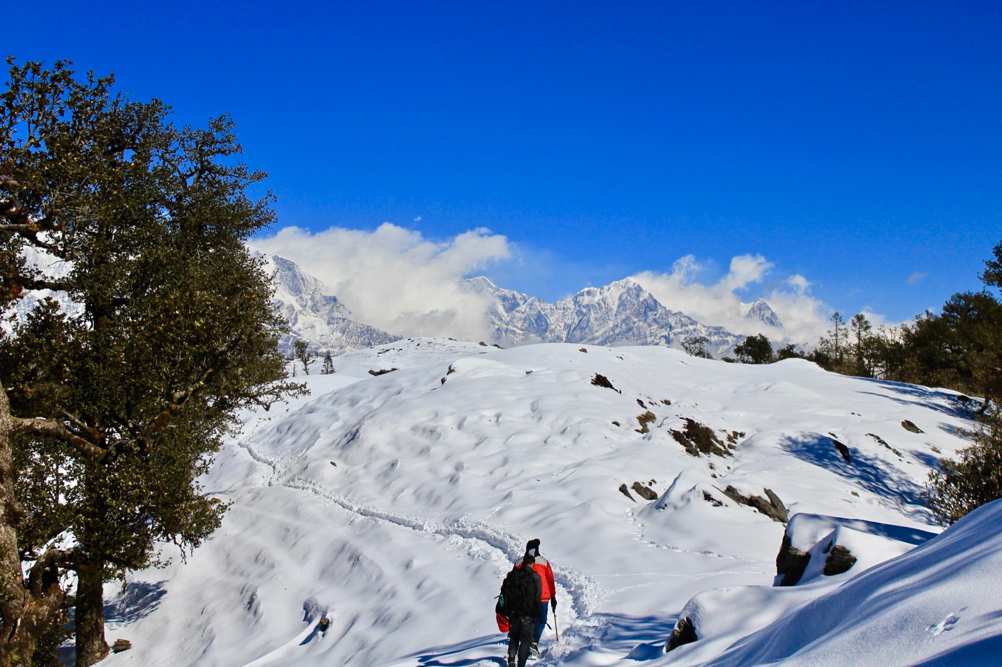 Trekkers crossing a snowy ridge towards Brahmatal below jagged peaks